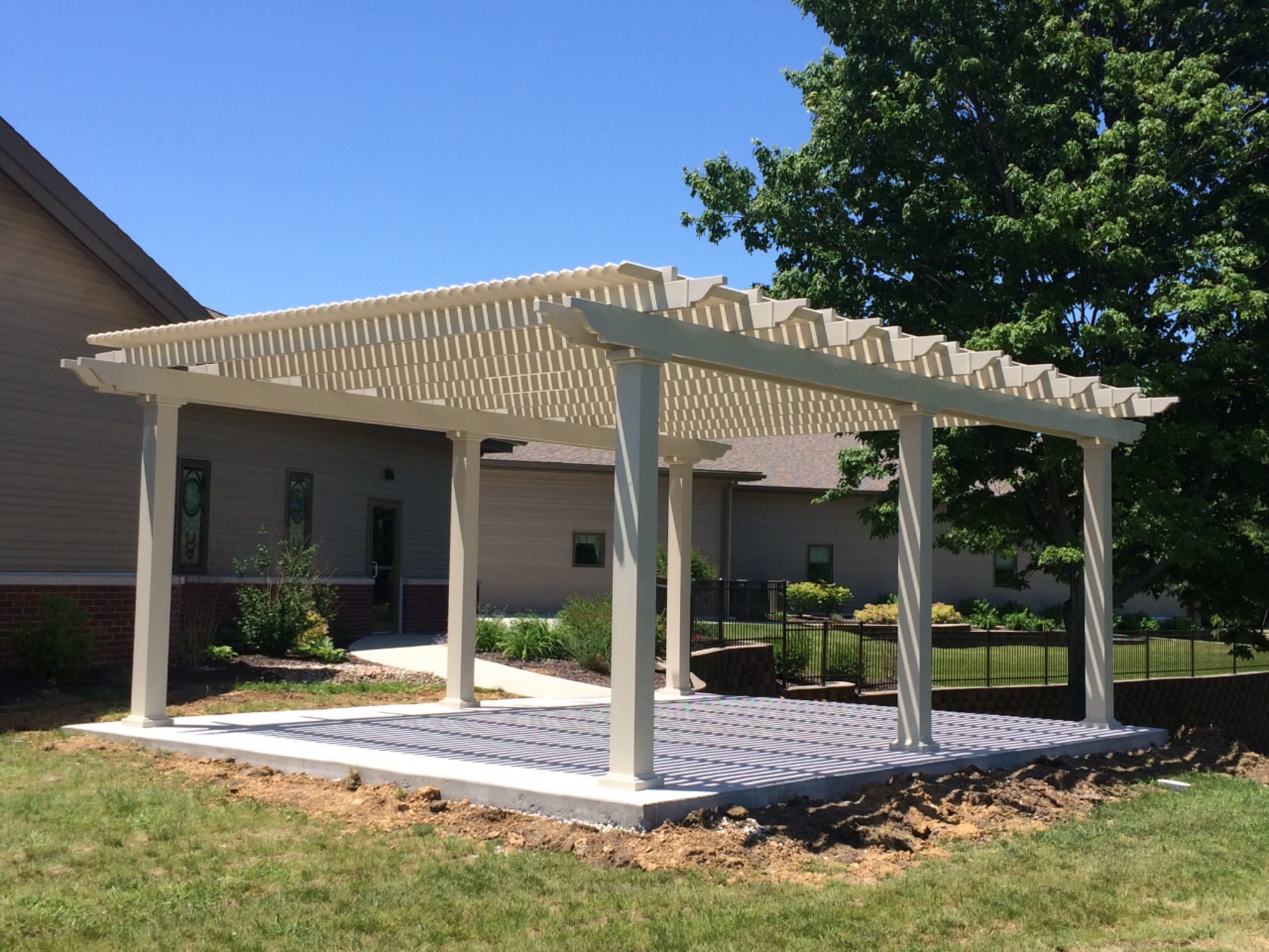 This white pergola is providing shade over an open, empty area. There is a house behind the pergola, and a sidewalk connecting the house to the pergola area.