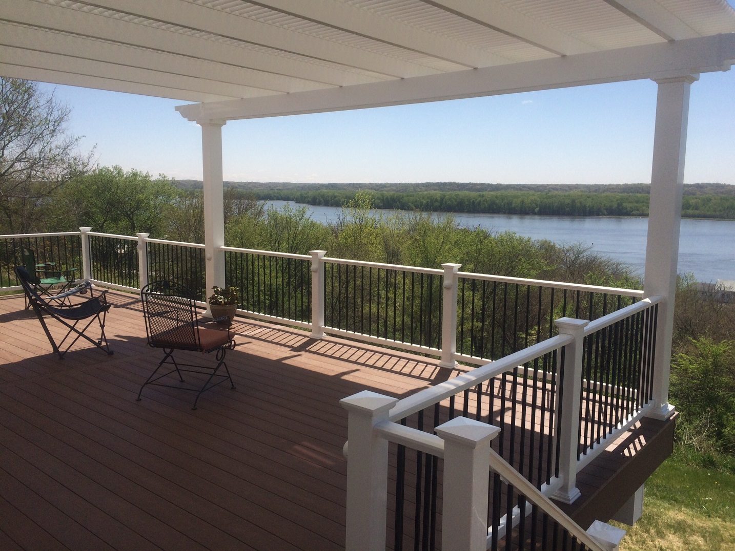 This large deck has a white pergola attached to provide shade. There are a couple chairs on the deck looking out to a body of water. There is a black and white railing across the deck’s perimeter.