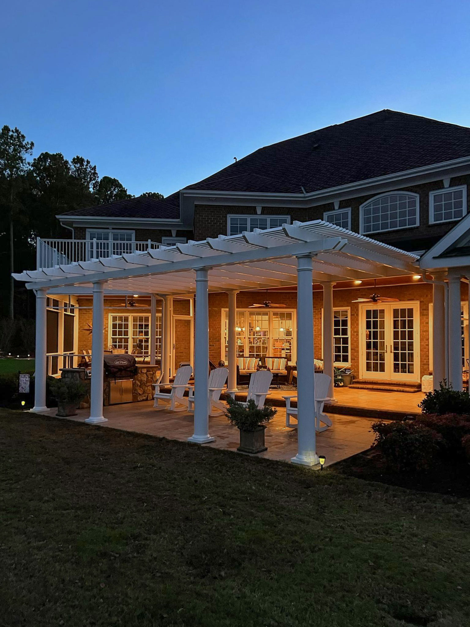 Large white traditional pergola with round post columns on a large traditional brick home at night
