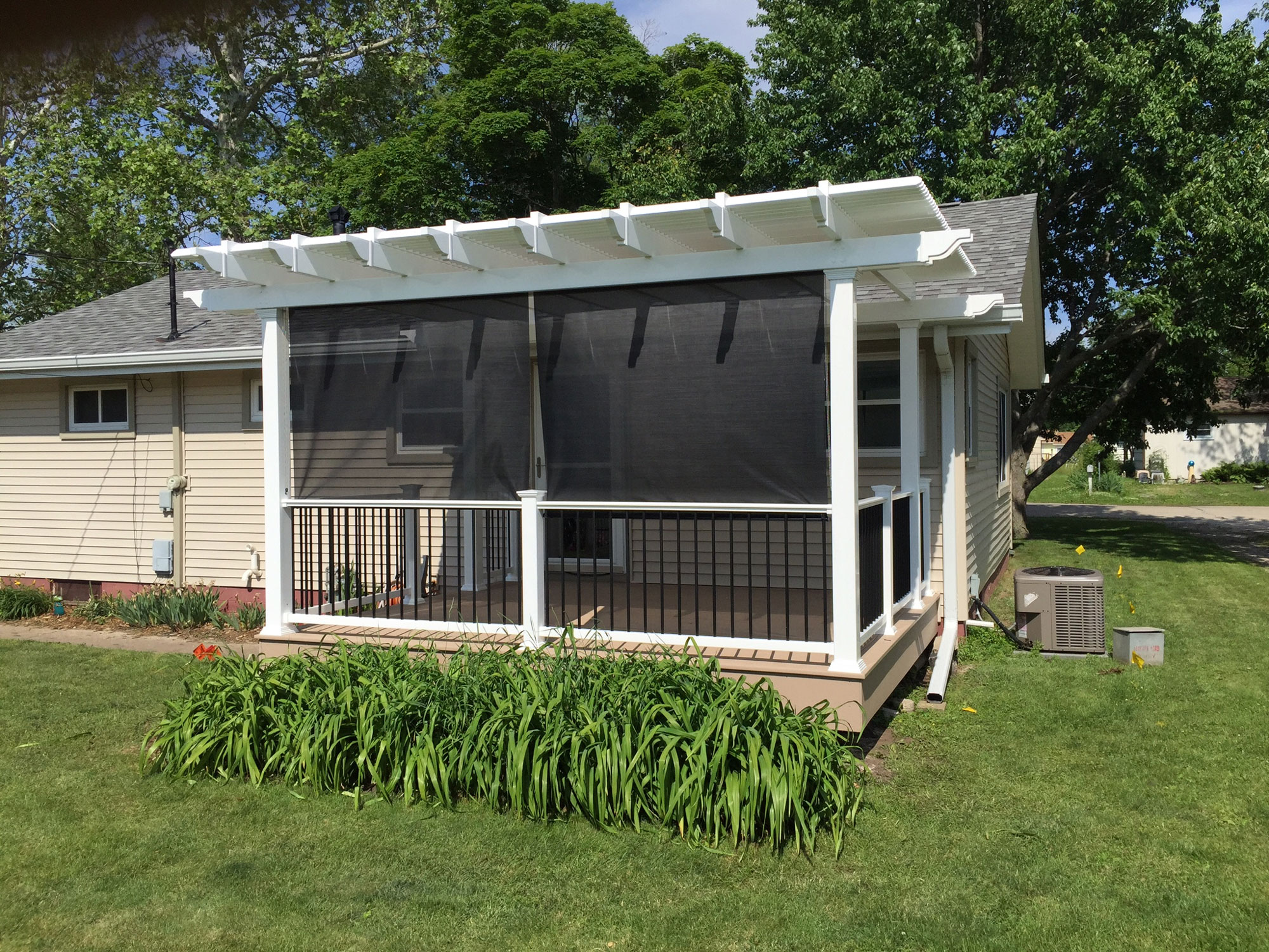 This small house extended its space by adding a back porch covered by a white pergola. They have leafy plants next to their brown porch and a sidewalk near it.