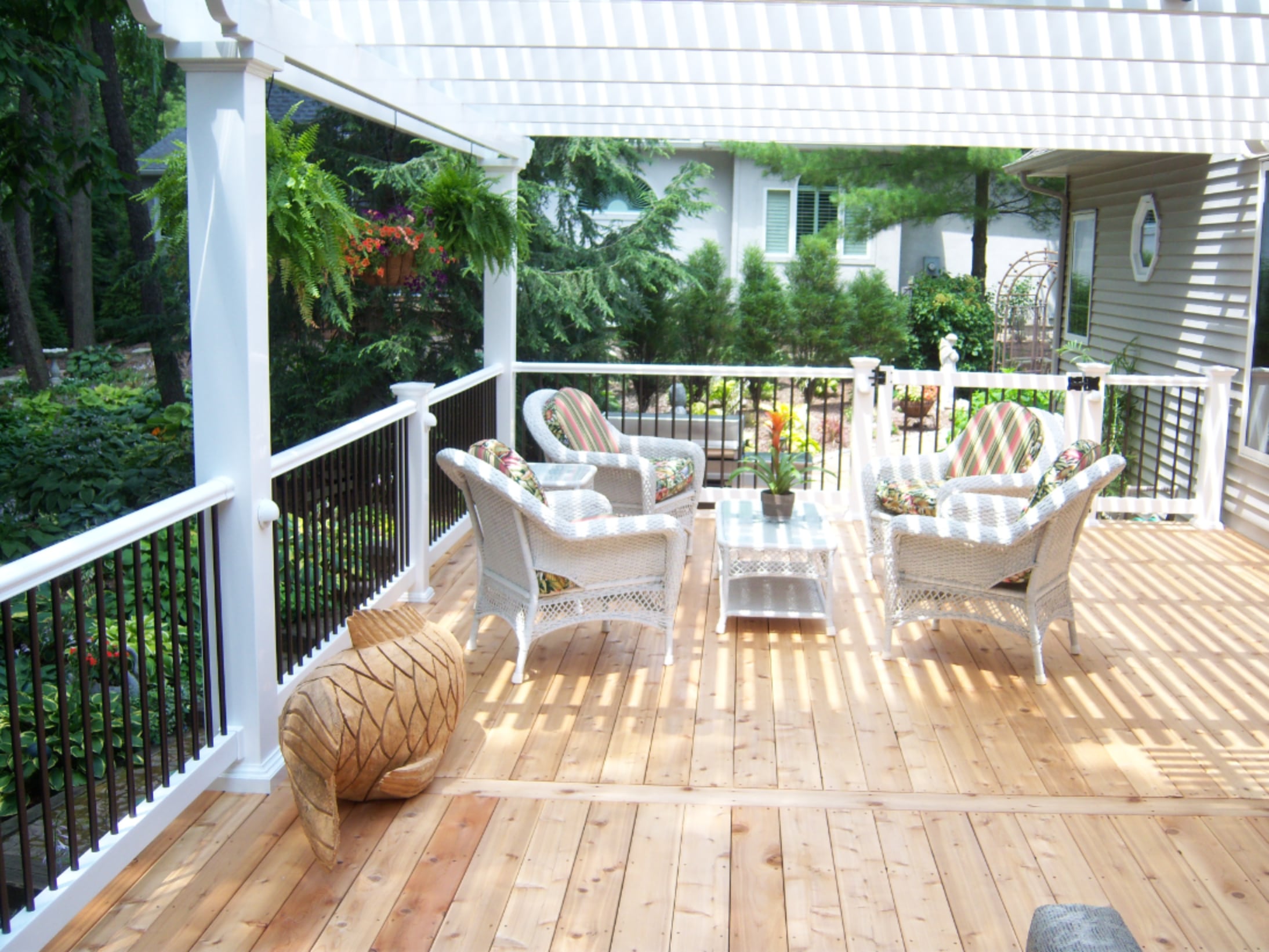 This porch has light wood floors, a white pergola and a white and black railing. There is a seating area with white wicker furniture and striped cushions.