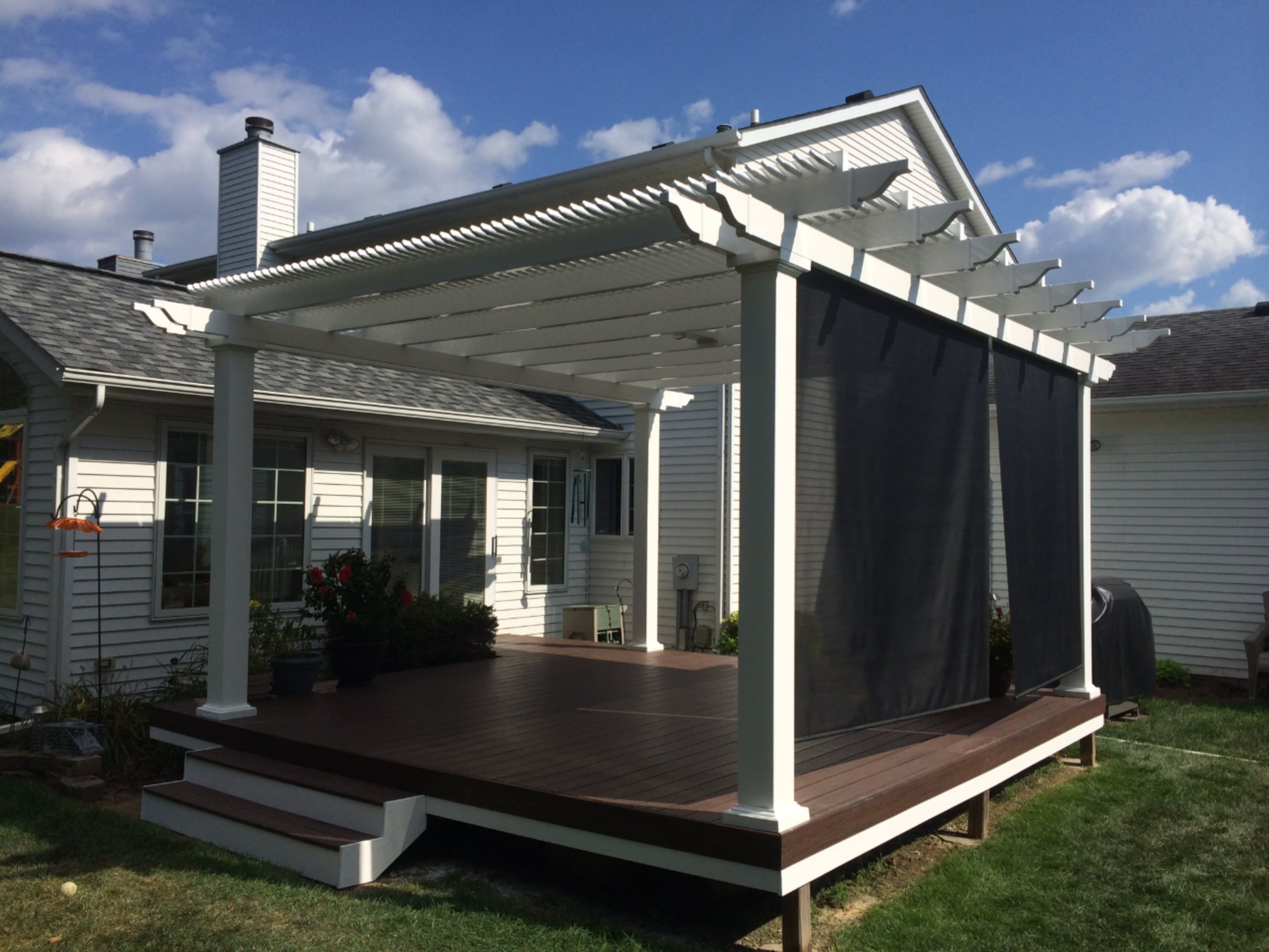 This home has a back decking area with a white pergola attached. There is a screen on the side of the pergola, and there is no furniture on the deck under the pergola.