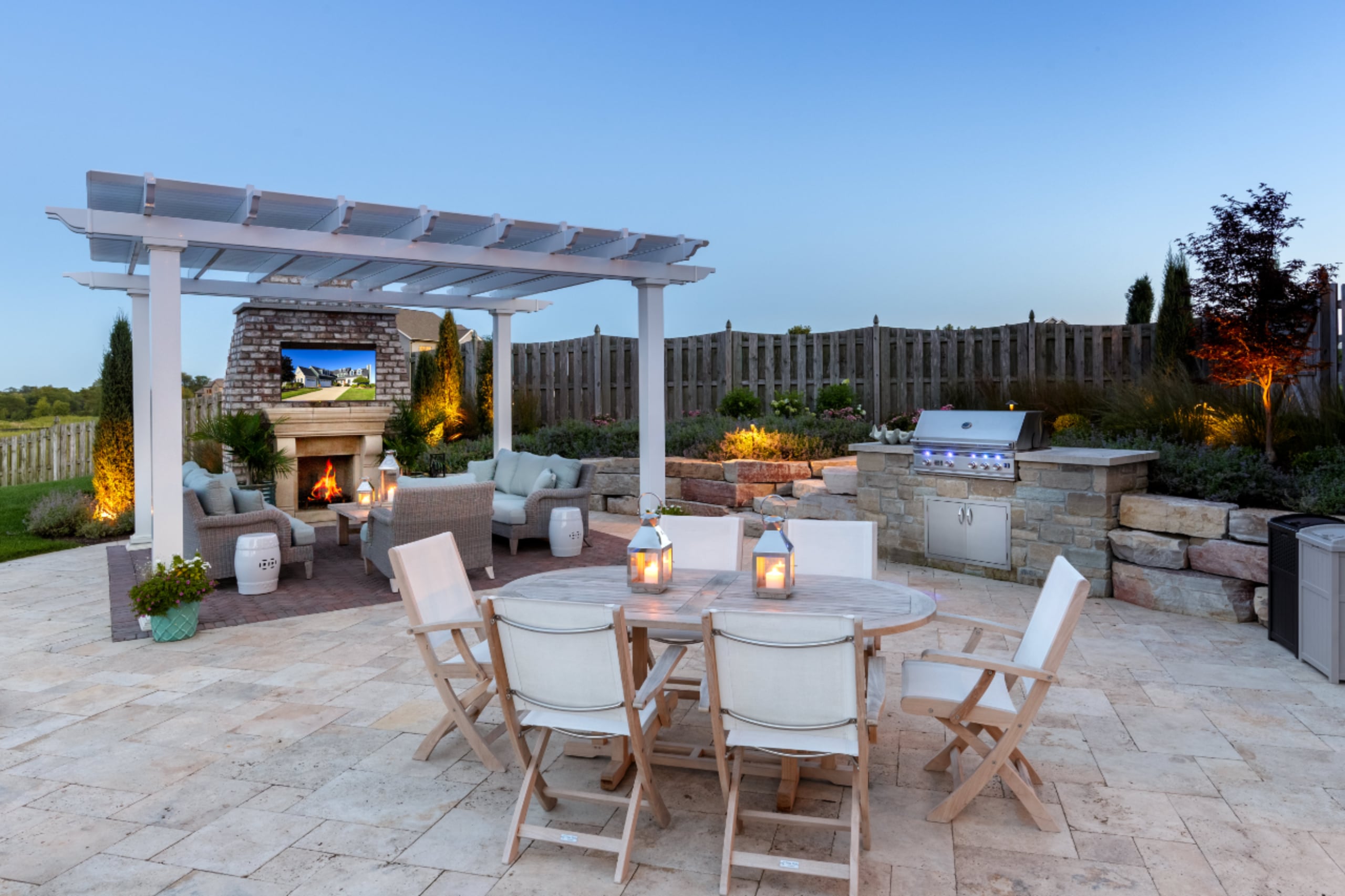 This outdoor space has a pergola covering a seating area with two couches, a chair, a fireplace and side tables. There is an eating area with six chairs and two lanterns on the table.