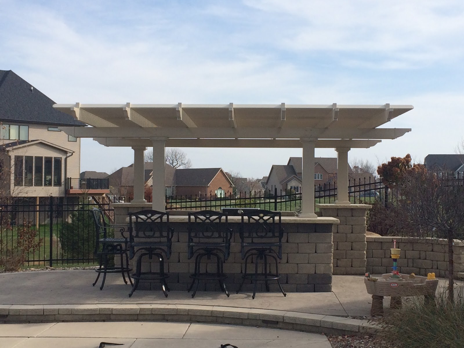 This outdoor bar and the cooking area have large concrete blocks and a tan pergola. There are black high chairs pulled up to the bar area.