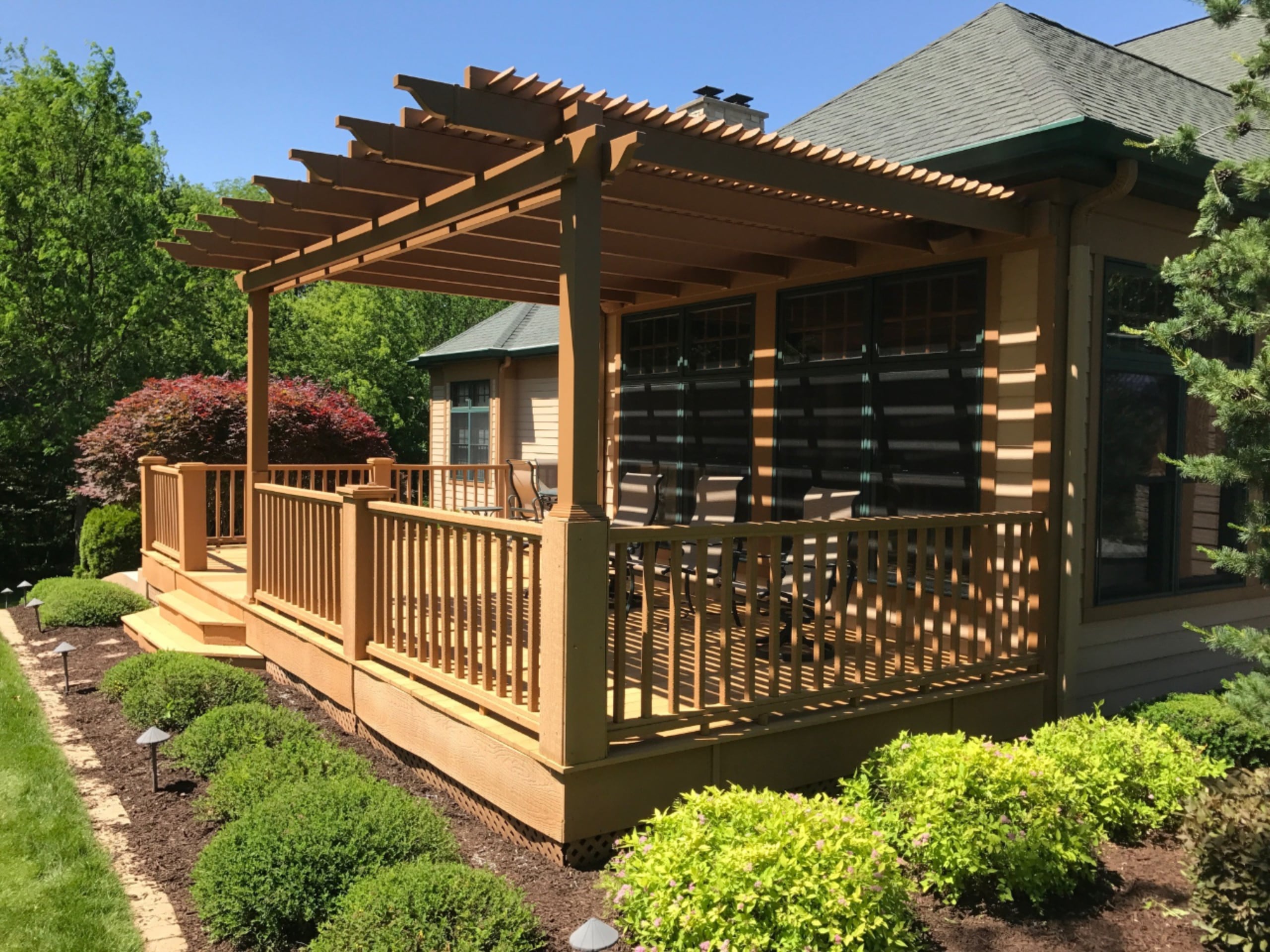 This brown house has a painted brown pergola covering half of the porch. There are green bushes around the porch with brown mulch.