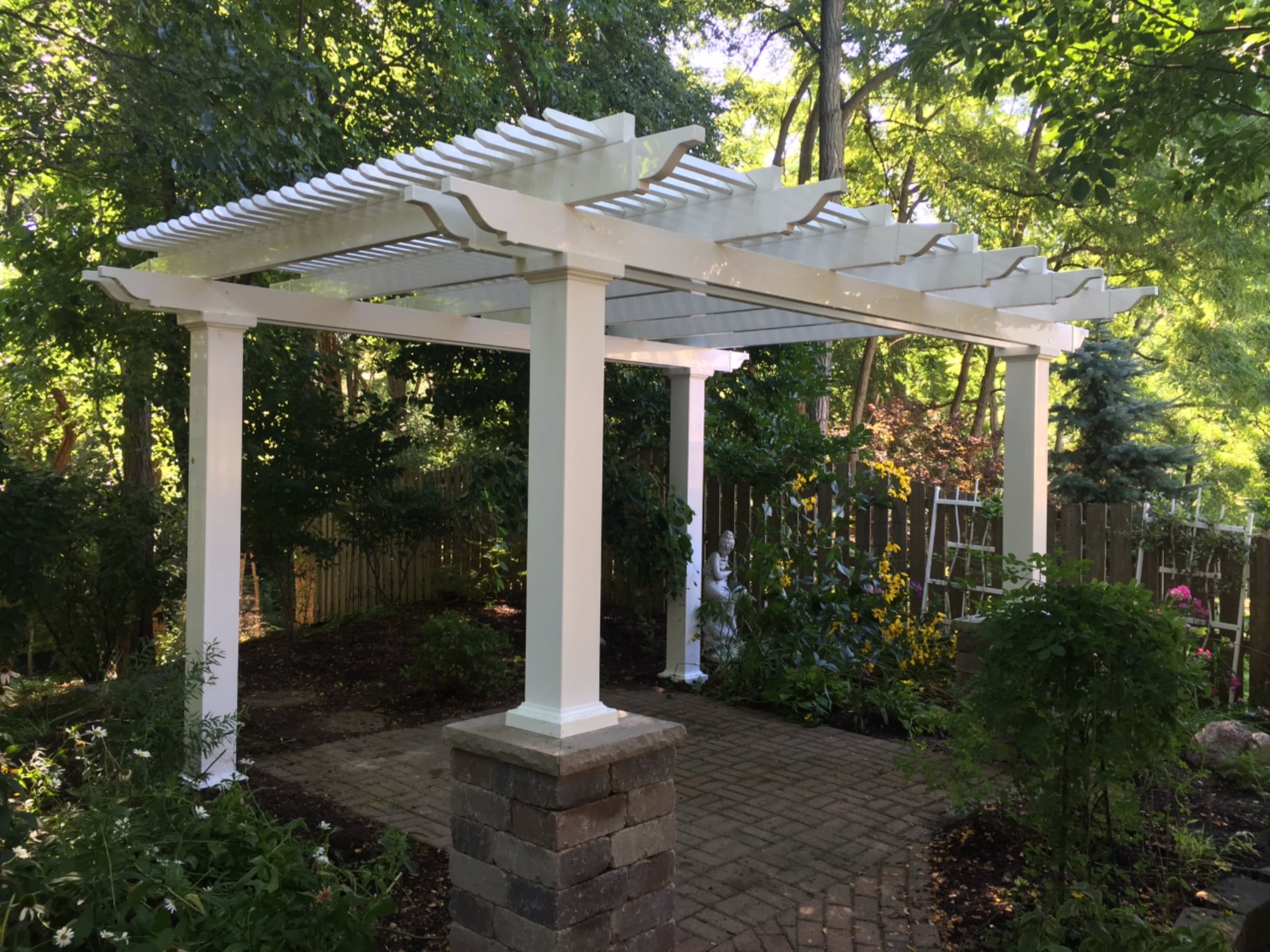 This white pergola is covering an open area with brick flooring. There are plants and trees all around the pergola.