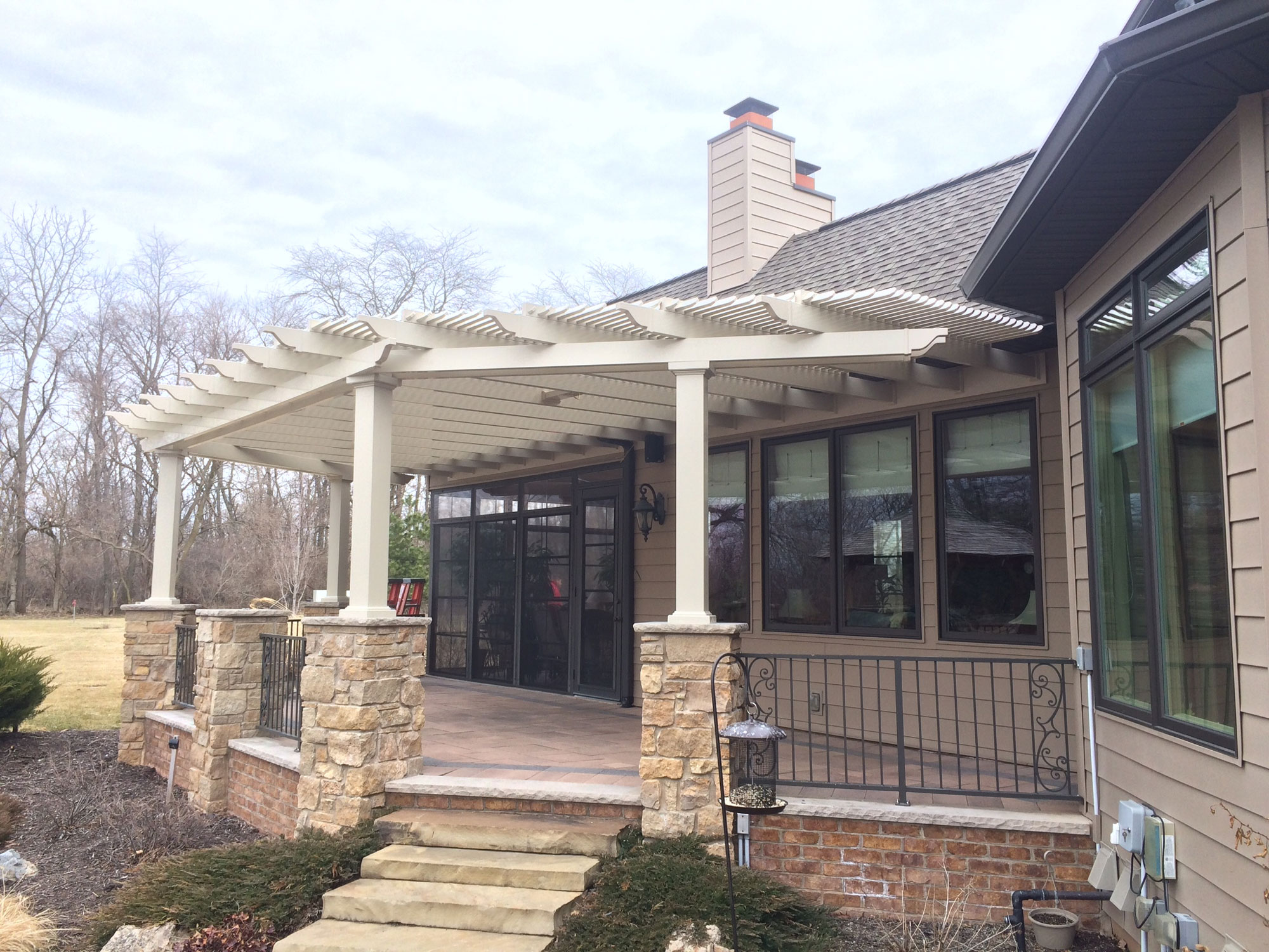 This brown home installed a tan pergola to cover their back porch. They have stone column connecting the pergola to the foundation, along with stone steps leading to the deck area.