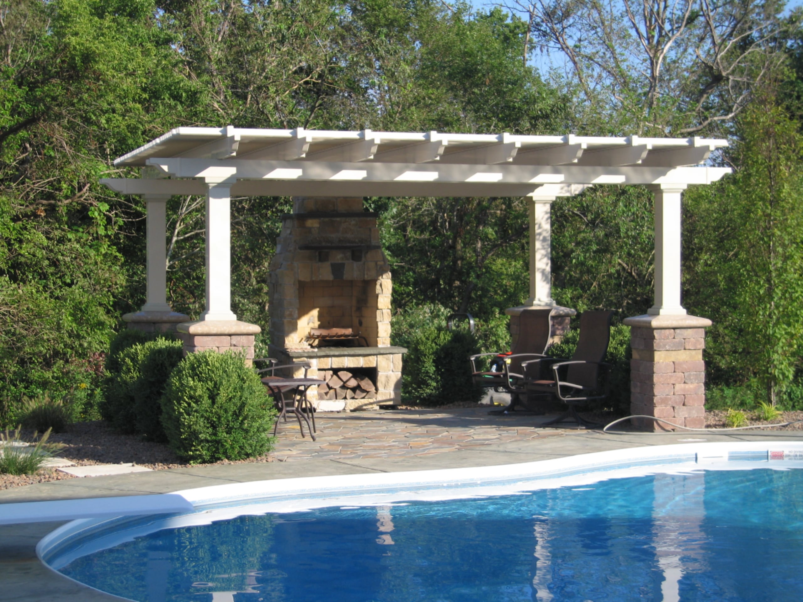 This white pergola is resting on brick pillars, covering an outdoor eating area next to an inground pool. There are trees behind the eating area and bushes next to the pillars.